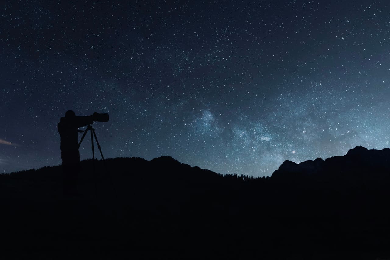 A person with a telescope silhouetted on a ridge under a starry sky